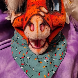 May include: A detailed close-up of a furry orange and white animal head with large blue eyes. The head is wearing a teal bandana with a pattern of Christmas lights. The background is a purple, textured fabric.