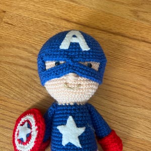 Op de afbeelding: Een handgemaakte Captain America pop, gehaakt in blauw, rood en wit garen. De pop heeft een blauw masker met een witte "A", een witte ster en een rood-wit schild. De pop is ongeveer 20 cm hoog.