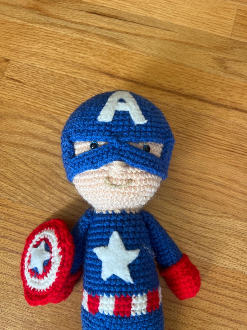 Op de afbeelding: Een handgemaakte Captain America pop, gehaakt in blauw, rood en wit garen. De pop heeft een blauw masker met een witte "A", een witte ster en een rood-wit schild. De pop is ongeveer 20 cm hoog.