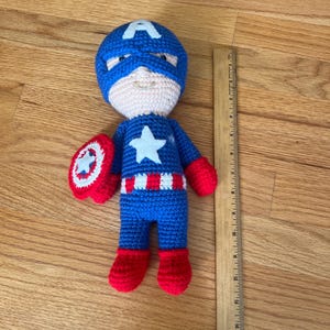 Op de afbeelding: Een handgemaakte Captain America amigurumi pop. De pop is voornamelijk blauw met rode en witte accenten, waaronder een ster op de borst en een schild. De pop is ongeveer 18 cm hoog.