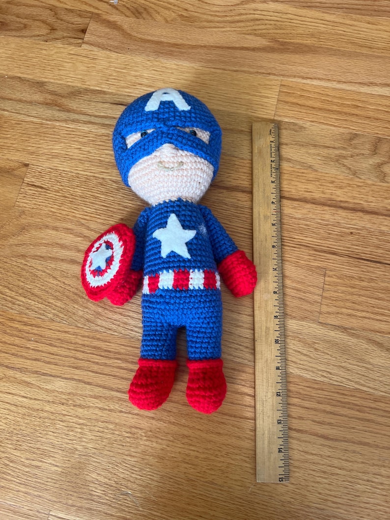 Op de afbeelding: Een handgemaakte Captain America amigurumi pop. De pop is voornamelijk blauw met rode en witte accenten, waaronder een ster op de borst en een schild. De pop is ongeveer 18 cm hoog.