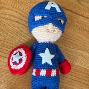 Op de afbeelding: Een handgemaakte gehaakte pop van Captain America. De pop is voornamelijk blauw met een witte ster op de borst en een witte "A" op het blauwe masker. De pop houdt een rood-wit schild vast met een witte ster.