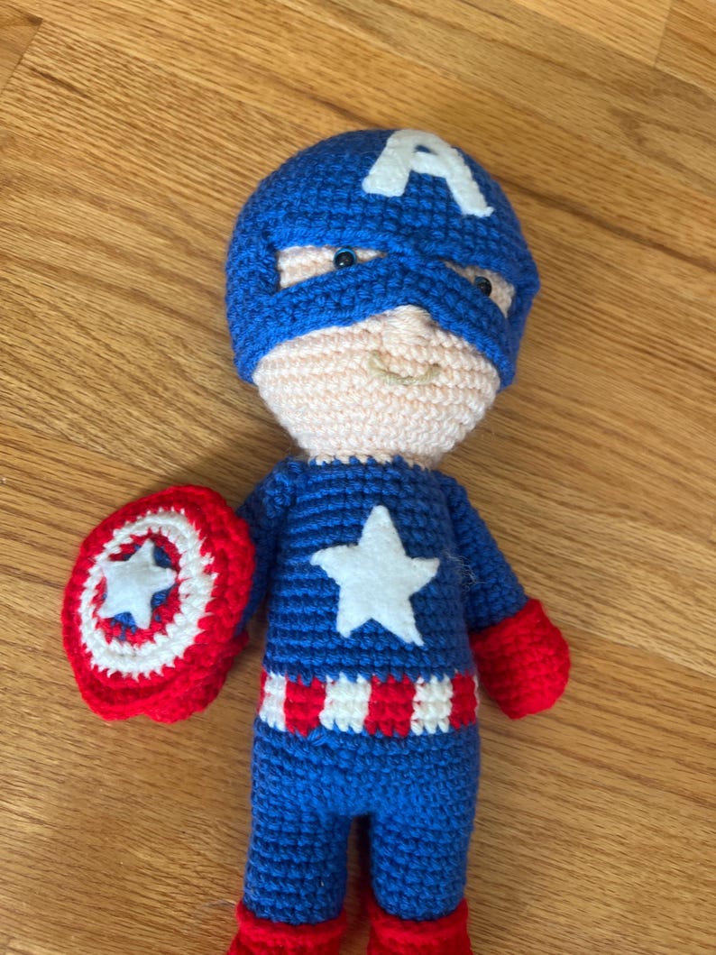 Op de afbeelding: Een handgemaakte gehaakte pop van Captain America. De pop is voornamelijk blauw met een witte ster op de borst en een witte "A" op het blauwe masker. De pop houdt een rood-wit schild vast met een witte ster.