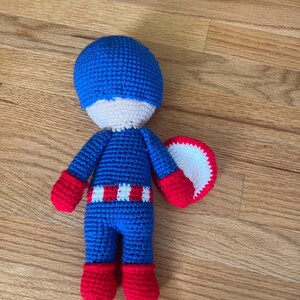 Op de afbeelding: Een handgemaakte gehaakte pop die Captain America voorstelt. De pop heeft een blauwe helm, uniform en laarzen, met rode handschoenen en een wit en rood schild. De pop is ongeveer 20 cm hoog.