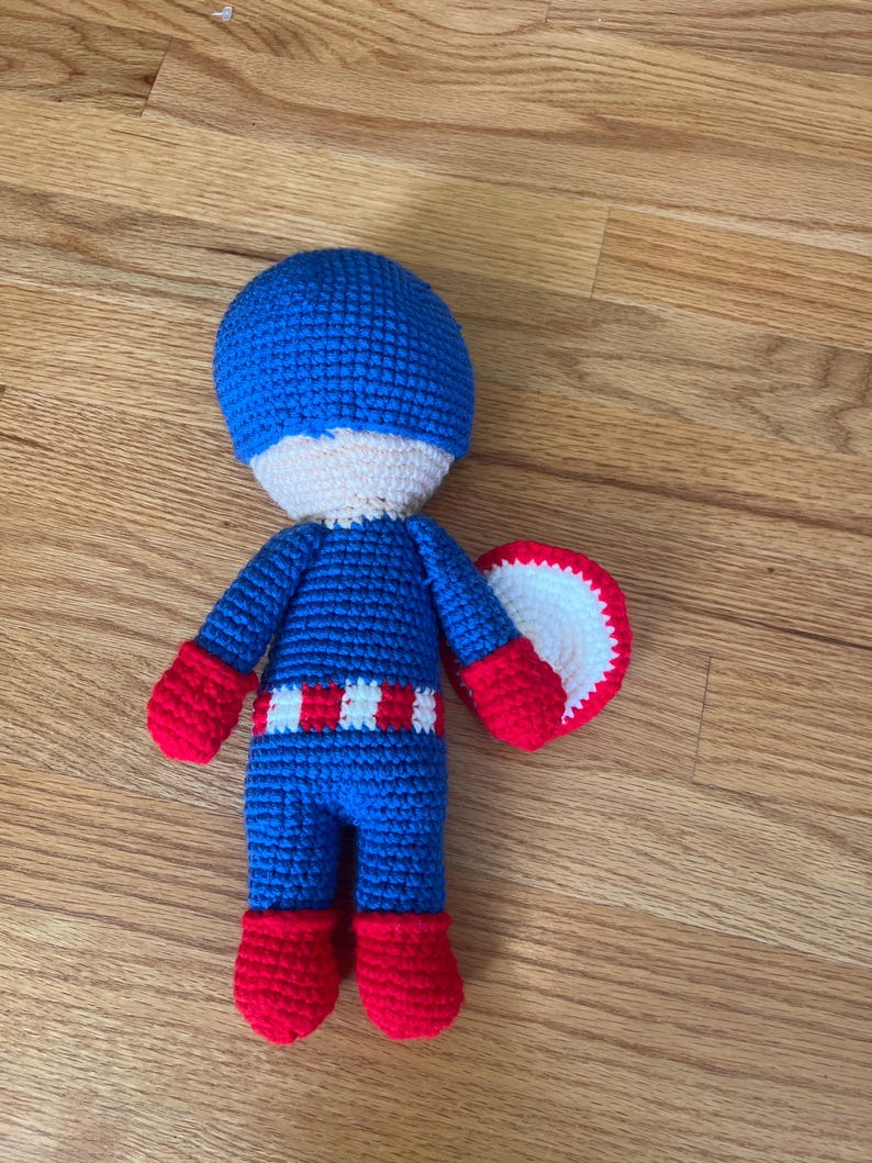 Op de afbeelding: Een handgemaakte gehaakte pop die Captain America voorstelt. De pop heeft een blauwe helm, uniform en laarzen, met rode handschoenen en een wit en rood schild. De pop is ongeveer 20 cm hoog.