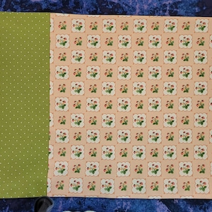 May include: A fabric panel with three sections. The left section is a light pink color, the middle section is a green color with white polka dots, and the right section is a peach color with a repeating pattern of white squares with green leaves and red berries.