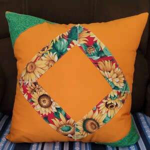 May include: Orange throw pillow with a diamond-shaped panel in the center. The panel is surrounded by a border of fabric with a sunflower pattern. The pillow is made of a soft, comfortable fabric.