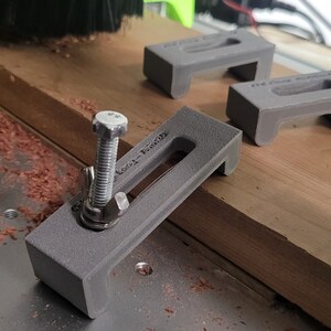 Puede incluir: Tres abrazaderas grises impresas en 3D con un tornillo roscado en una de las abrazaderas. Las abrazaderas est&aacute;n sobre una superficie de madera con virutas de madera.