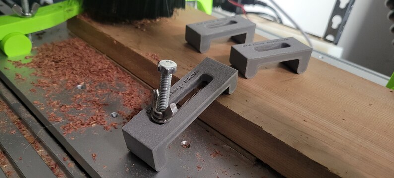 Puede incluir: Tres abrazaderas grises impresas en 3D con un tornillo roscado en una de las abrazaderas. Las abrazaderas est&aacute;n sobre una superficie de madera con virutas de madera.
