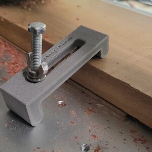 Puede incluir: Una abrazadera gris impresa en 3D con un tornillo roscado y una tuerca de metal. La abrazadera est&aacute; dise&ntilde;ada para sujetar la madera en su lugar mientras se trabaja en ella. La abrazadera est&aacute; sentada sobre una superficie de madera.