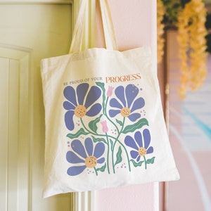 Puede incluir: Una bolsa de lona blanca con un diseño floral y el texto "Esté orgulloso de su progreso". Las flores son azules, verdes y rosas.