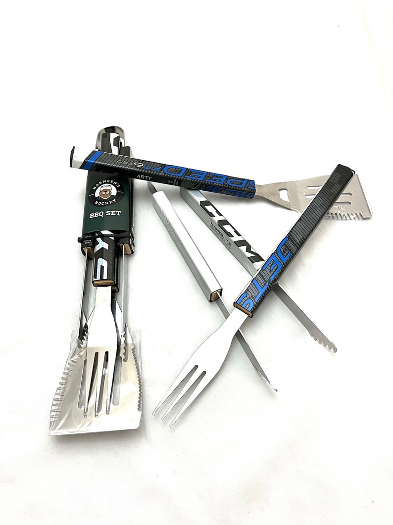 K&ouml;nnte beinhalten: Ein Set aus vier Grillwerkzeugen mit Hockeyschl&auml;gergriffen. Die Werkzeuge umfassen einen Spatel, eine Zange, eine Gabel und eine Grillb&uuml;rste. Die Griffe sind schwarz und blau mit wei&szlig;en Buchstaben. Die Werkzeuge sind in einer durchsichtigen Plastikt&uuml;te mit einem Etikett mit der Aufschrift "BBQ Set" verpackt.