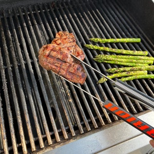 K&ouml;nnte beinhalten: Nahaufnahme eines Steaks, das auf einem schwarzen Grill mit Spargel an der Seite gegrillt wird. Das Steak wird mit einer roten und schwarzen Zange gewendet.