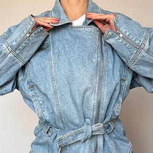 Puede incluir: Chaqueta vaquera azul claro con cremallera frontal, mangas largas y cintur&oacute;n en la cintura. La chaqueta tiene un cuello cl&aacute;sico y detalles de cremallera en las mangas. El tejido vaquero tiene un lavado descolorido.