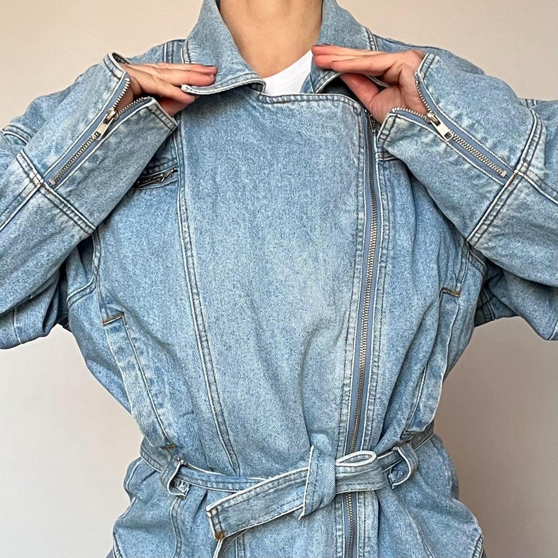 Puede incluir: Chaqueta vaquera azul claro con cremallera frontal, mangas largas y cintur&oacute;n en la cintura. La chaqueta tiene un cuello cl&aacute;sico y detalles de cremallera en las mangas. El tejido vaquero tiene un lavado descolorido.