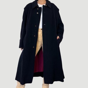 Peut inclure: Long manteau noir en laine avec une fermeture boutonnée et un col. Le manteau est long et doublé de rouge au niveau de l'ourlet. Il est porté sur une chemise blanche et un pantalon beige.