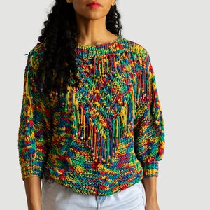 May include: A colourful knitted jumper with a rainbow pattern and fringe details. The jumper has a round neckline and long sleeves.