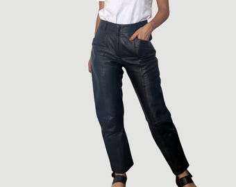 May include: Black leather pants with a high waist and a straight leg. The pants have pockets and a button and zipper closure. The pants are worn with a white top and black sandals.