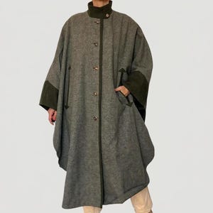 May include: A long, grey wool-blend coat featuring a dark olive green stand-up collar and matching dark olive green sleeve cuffs. The coat has a central button-down closure with visible buttons and two vertical slit pockets on the front panels.