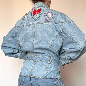 Puede incluir: Chaqueta vaquera de lavado claro con parches bordados de coraz&oacute;n y flores en la espalda. La chaqueta presenta una cintura con cintur&oacute;n, cierre de cremallera y cuello alto. El bordado incluye un lazo rojo y varios dise&ntilde;os florales coloridos.