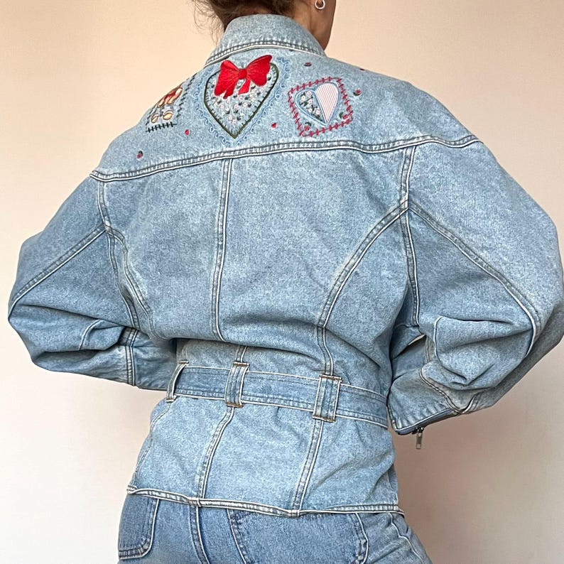 Puede incluir: Chaqueta vaquera de lavado claro con parches bordados de coraz&oacute;n y flores en la espalda. La chaqueta presenta una cintura con cintur&oacute;n, cierre de cremallera y cuello alto. El bordado incluye un lazo rojo y varios dise&ntilde;os florales coloridos.