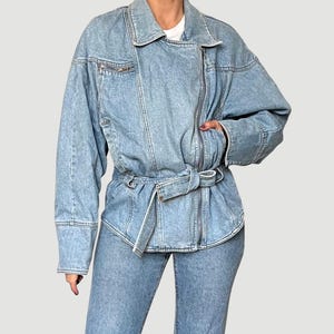 Puede incluir: Chaqueta vaquera azul claro con cierre de cremallera y cintur&oacute;n en la cintura. La chaqueta tiene mangas largas y un cuello cl&aacute;sico. El modelo lleva vaqueros vaqueros a juego. El estilo general es casual y moderno.