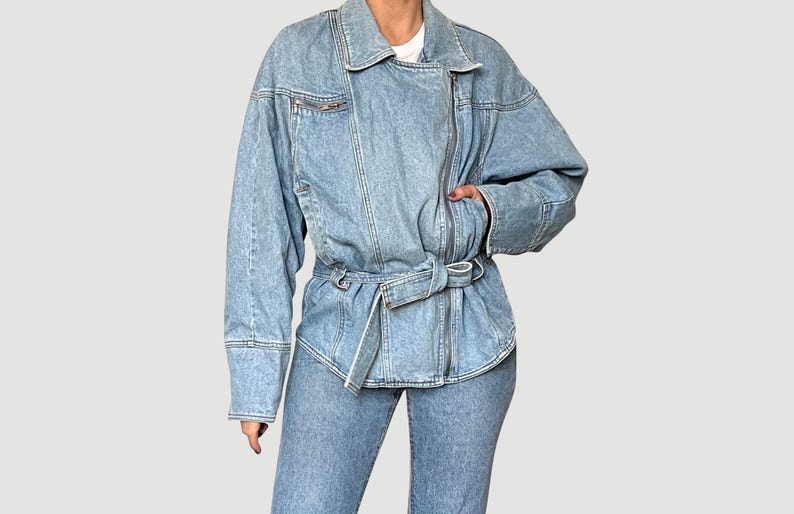 Puede incluir: Chaqueta vaquera azul claro con cierre de cremallera y cintur&oacute;n en la cintura. La chaqueta tiene mangas largas y un cuello cl&aacute;sico. El modelo lleva vaqueros vaqueros a juego. El estilo general es casual y moderno.
