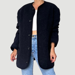 Puede incluir: Chaqueta abotonada azul marino oscuro con cuello redondo. La chaqueta se lleva sobre una camiseta blanca y vaqueros azules. La chaqueta tiene un aspecto texturizado y un diseño clásico.
