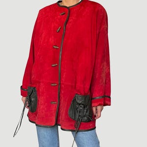 May include: A red suede jacket with black leather trim and pockets. The jacket features wooden toggle closures and black leather pockets with drawstring details. The jacket is a long, oversized style.
