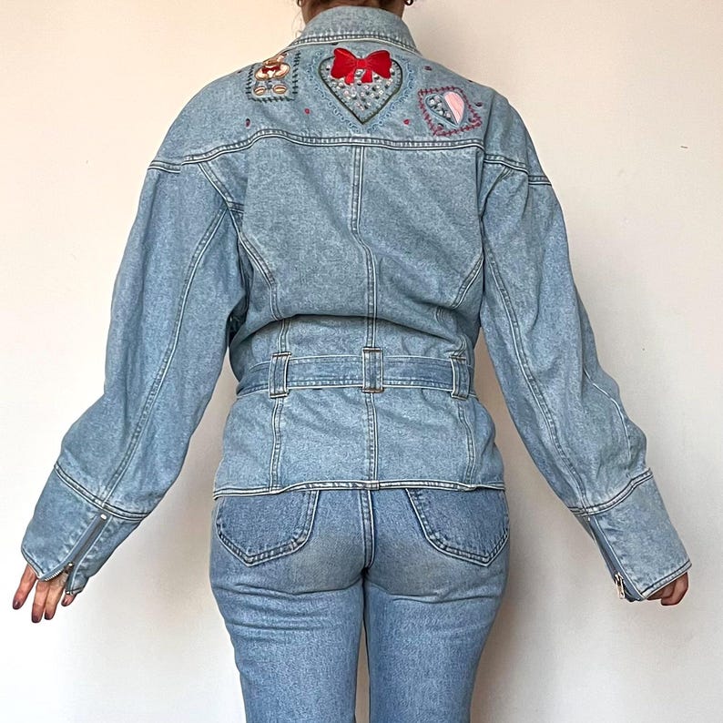 Puede incluir: Chaqueta vaquera azul claro con detalles bordados de coraz&oacute;n y lazo en la espalda. La chaqueta tiene cintura con cintur&oacute;n y mangas largas con pu&ntilde;os con cremallera. El modelo tambi&eacute;n lleva vaqueros azules claros.