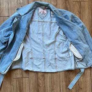 Puede incluir: Chaqueta vaquera azul claro con un dise&ntilde;o de coraz&oacute;n bordado en la espalda. La chaqueta tiene cuello, mangas largas y cintur&oacute;n. El forro interior es blanco.