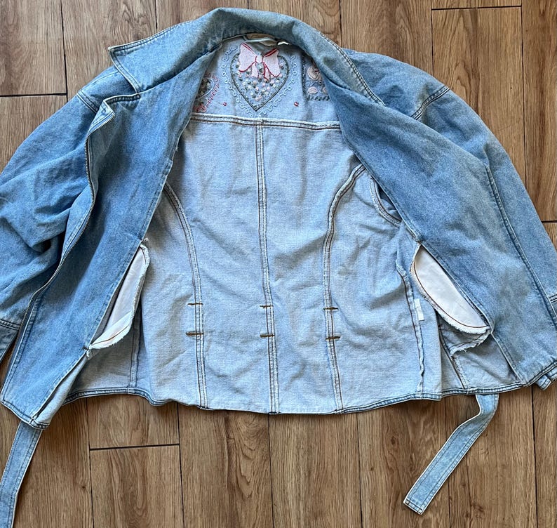 Puede incluir: Chaqueta vaquera azul claro con un dise&ntilde;o de coraz&oacute;n bordado en la espalda. La chaqueta tiene cuello, mangas largas y cintur&oacute;n. El forro interior es blanco.