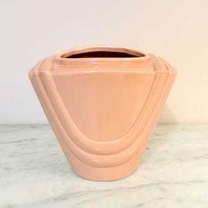 May include: A peach-colored ceramic vase with a geometric design. The vase has a wide, open top and a series of curved, recessed lines on the lower portion. The vase is sitting on a marble surface.