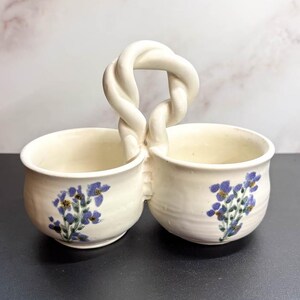 May include: Cream-colored ceramic serving dish with two bowls connected by a twisted handle. Each bowl is decorated with a blue floral design. The dish is on a dark surface.
