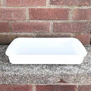May include: A rectangular, white plastic tray with rounded corners. The tray is empty and sits on a concrete surface in front of a red brick wall. The tray is likely used for food preparation or storage.
