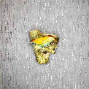May include: A ceramic bird figurine perched on a tree branch. The bird has yellow, blue, and red coloring. The tree branch and base are green and brown. The figurine is a decorative item.