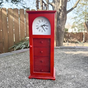 May include: A red wooden clock with a white clock face featuring black Roman numerals. The clock has a small cabinet with three drawers. The clock is outdoors, set on a concrete surface.