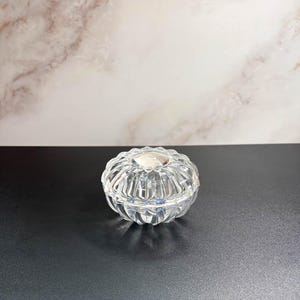 May include: A clear, round, glass trinket box with a lid. The box has a ribbed design and a small, silver-colored accent on the lid. The background is a marble pattern.