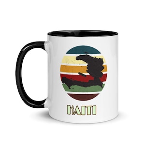 Może przedstawiać: Biały kubek ceramiczny z czarnym rantem. Kubek przedstawia grafikę w stylu retro wyspy Haiti z kolorowym, paskowanym słońcem z tyłu. Tekst "HAITI" jest nadrukowany na zielono pod grafiką.