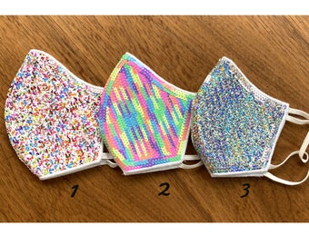 May include: Three face masks with different sequin patterns. The first mask has a white background with multi-colored sequins. The second mask has a white background with a rainbow stripe pattern. The third mask has a white background with silver sequins.