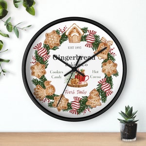 May include: Round wall clock with a black frame featuring a gingerbread theme. The clock face has a white background with a wreath of gingerbread cookies, candy, and greenery. Text reads "Gingerbread Baking Co." and "Fresh Daily."