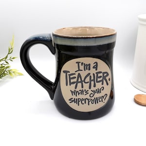 May include: Black ceramic mug with a large handle, featuring the text "I'm a TEACHER. What's your superpower?" in a playful font. The mug has a beige interior and a dark blue rim. A white sugar container and wooden spoon are in the background.