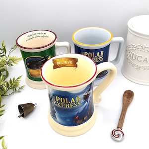 May include: Three ceramic mugs with handles, featuring "Polar Express" imagery and text. One mug has a green exterior with the quote "The bell still rings for me, as it does for all who truly believe." A small bell and a wooden spoon with a candy cane design are also present.