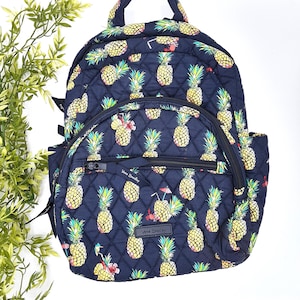 May include: Navy blue quilted backpack with a repeating pineapple pattern in yellow, green, and red. The backpack has a top handle, a front zippered pocket, and side pockets. The brand name "Vera Bradley" is visible on the front.