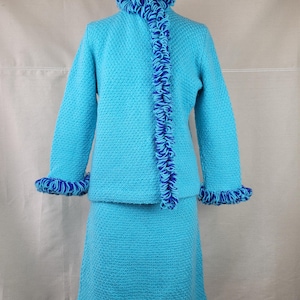 May include: A turquoise knitted sweater and skirt set with blue and turquoise trim. The sweater has a ruffled collar and cuffs. The skirt is a-line and hits above the knee.