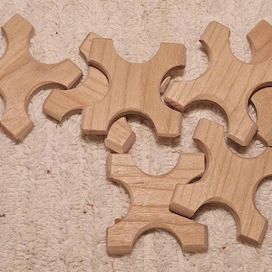 May include: Several unfinished wooden puzzle pieces with a cross-like shape are scattered on a white, textured surface. The pieces are light brown and appear to be made of wood, with rounded edges and a smooth finish.