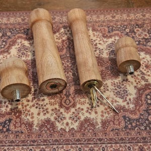 May include: Two cylindrical light brown wooden handles with rounded ends, accompanied by two matching wooden caps. One handle features a metal insert with a small metal tool protruding from the center, while the other shows a recessed metal fitting.