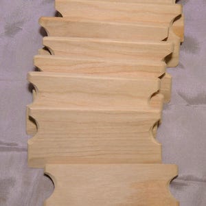 Puede incluir: Un conjunto de ocho placas de madera sin terminar con bordes redondeados y un recorte en cada extremo. Las placas están apiladas una encima de la otra.