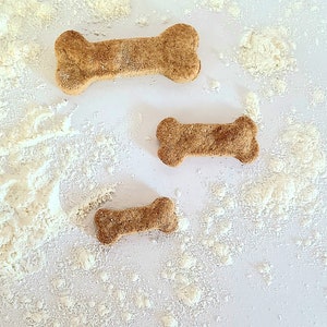 May include: Three dog bone-shaped treats on a white surface covered in white powder. The treats are different sizes and are a light brown color.