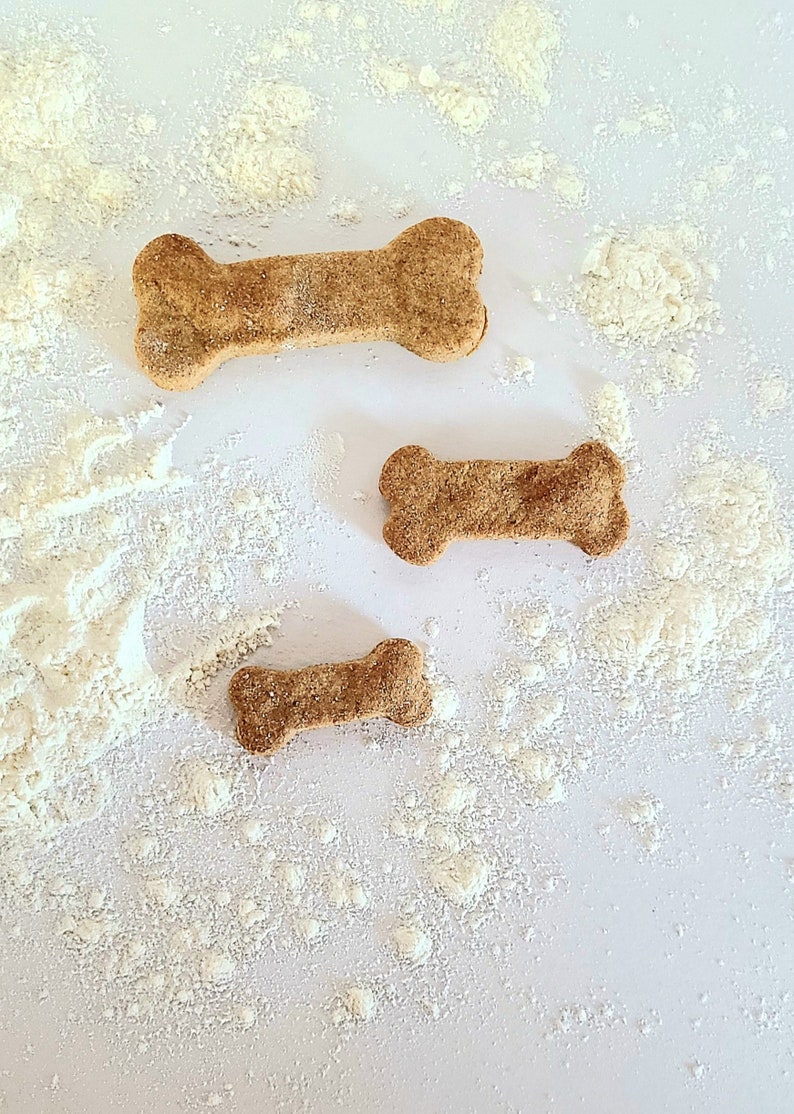 May include: Three dog bone-shaped treats on a white surface covered in white powder. The treats are different sizes and are a light brown color.